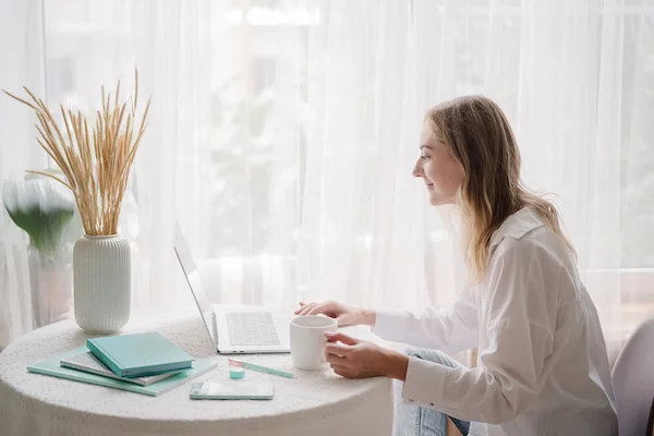 A women in white sits at a table with a hot drink in a mug looking at a computer.