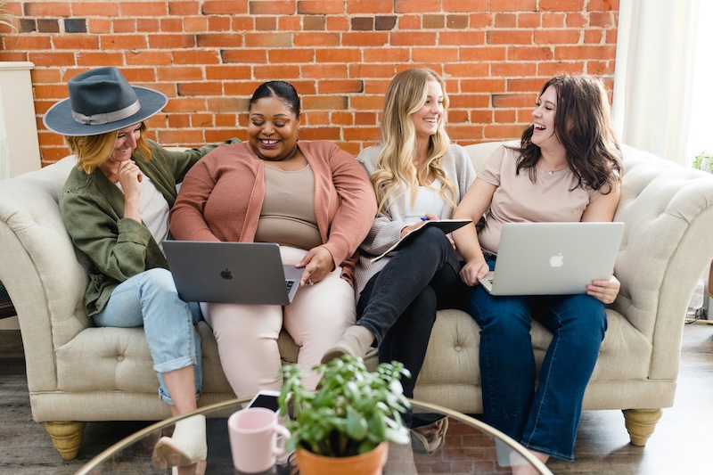 four women on a couch talking while looking at computers